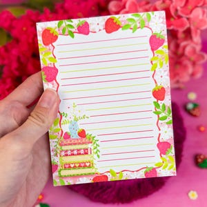 May include: A lined notepad with a strawberry and cake design. The notepad has a border of strawberries and green leaves. A small cake illustration is in the bottom left corner. The background is pink.