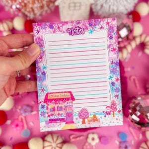 May include: A notepad with the word "Notes" at the top, featuring a pink and purple floral border. The notepad has lined pages and a drawing of a pink building with the text "Romance Bookstore". The background is pink with holiday decorations.