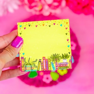 May include: A yellow sticky note with a decorative border of hearts and dots. The bottom of the note features a colorful illustration of books and potted plants. The note is held against a bright pink background.