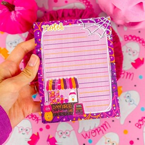 May include: A purple and pink notepad with the word "notes" at the top. The notepad has lined pages and a decorative border with a silver spiderweb design. The bottom of the notepad features a colorful cart with the text "BOOnards Pumpkinber cart".