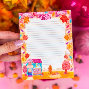 Könnte beinhalten: Ein farbenfroher Notizblock mit dem Wort "Notes" oben. Der Notizblock zeigt eine herbstliche Illustration mit einem kleinen Gebäude, Bäumen und einer Bank. Der Hintergrund ist rosa mit Herbstdekorationen.