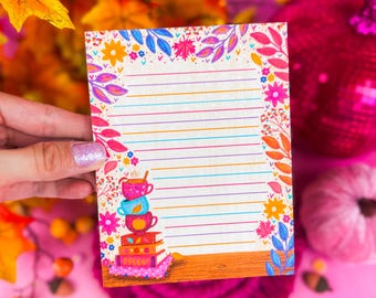 May include: A colorful notepad with a floral border and lined pages. The notepad features a stack of teacups and books illustration. The background includes autumn leaves, pumpkins, and decorative elements, all set against a pink backdrop.