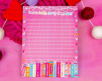 May include: Pink notepad with a glitter finish and the text "romance books are my happy place". The notepad has a lined interior and a bottom border of colorful books with titles like "One Bed", "Fated Mates", and "Enemies to Lovers".