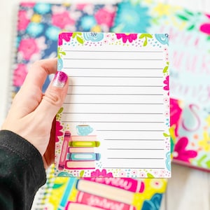 Puede incluir: Un bloc de notas floral rosa y azul con papel rayado. El bloc de notas tiene una pila de libros y una taza de café en la esquina. El bloc de notas se sostiene en una mano.