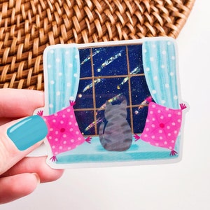 May include: A gray cat sits on a windowsill looking out at a starry night sky. The window is framed by blue curtains with white polka dots. Two pink pillows with white polka dots are on either side of the cat.