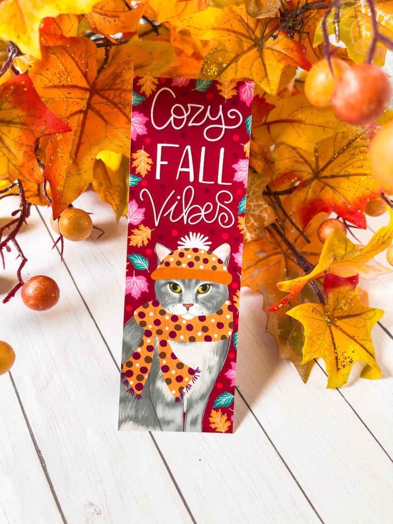 May include: A red bookmark with a gray cat wearing a brown and orange scarf and a hat. The bookmark has the text "Cozy Fall Vibes" in white.