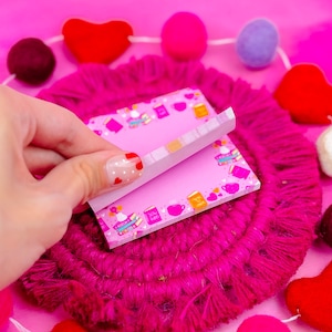 May include: Pink sticky notes with a book and coffee cup pattern. The notes are on a pink and white fuzzy surface with a string of pink, red, and purple felt balls.