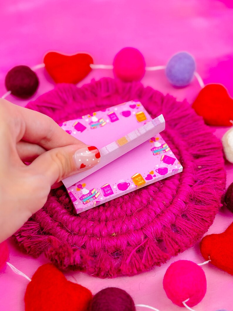May include: Pink sticky notes with a book and coffee cup pattern. The notes are on a pink and white fuzzy surface with a string of pink, red, and purple felt balls.
