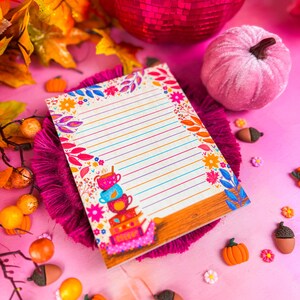 May include: A decorative notepad with colorful floral and leaf border. The notepad has lined pages and a stack of teacups illustration. The image includes a pink disco ball, a pink velvet pumpkin, and fall-themed decorations.