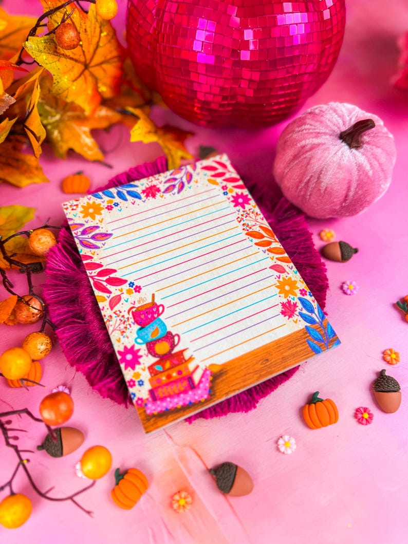 May include: A decorative notepad with colorful floral and leaf border. The notepad has lined pages and a stack of teacups illustration. The image includes a pink disco ball, a pink velvet pumpkin, and fall-themed decorations.