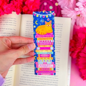 May include: A blue bookmark with a stack of colorful books and a cute orange cat sleeping on top. The bookmark has a starry night sky background with white stars and a string of white lights.