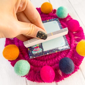 May include: A small notepad with the text "book club" is held over a bright pink, fringed coaster with colorful felt balls. The notepad has a blue background with stars and a stack of books. The felt balls are orange, teal, pink, yellow, and navy.