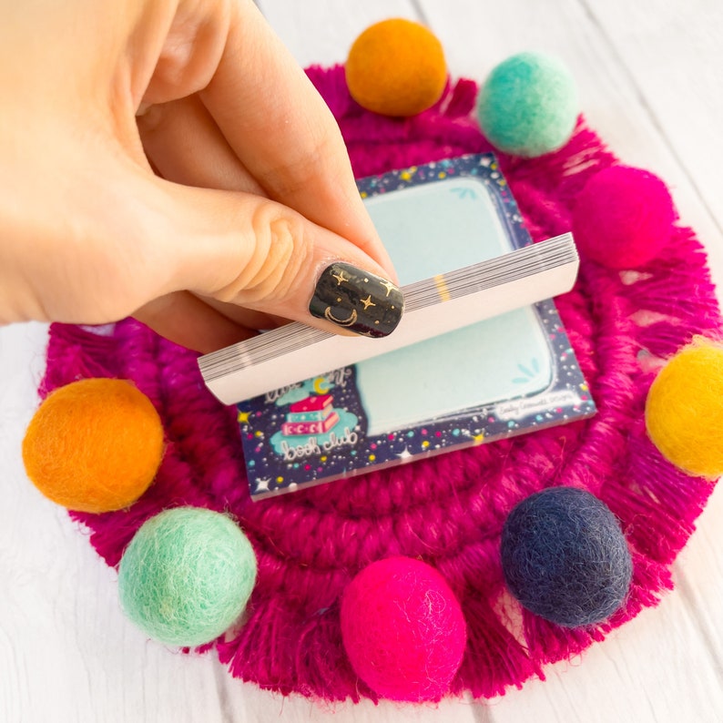 May include: A small notepad with the text "book club" is held over a bright pink, fringed coaster with colorful felt balls. The notepad has a blue background with stars and a stack of books. The felt balls are orange, teal, pink, yellow, and navy.