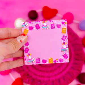 May include: Pink notepad with a border of colorful illustrations of books, coffee cups, and hearts. The notepad has a blank space in the center for writing.