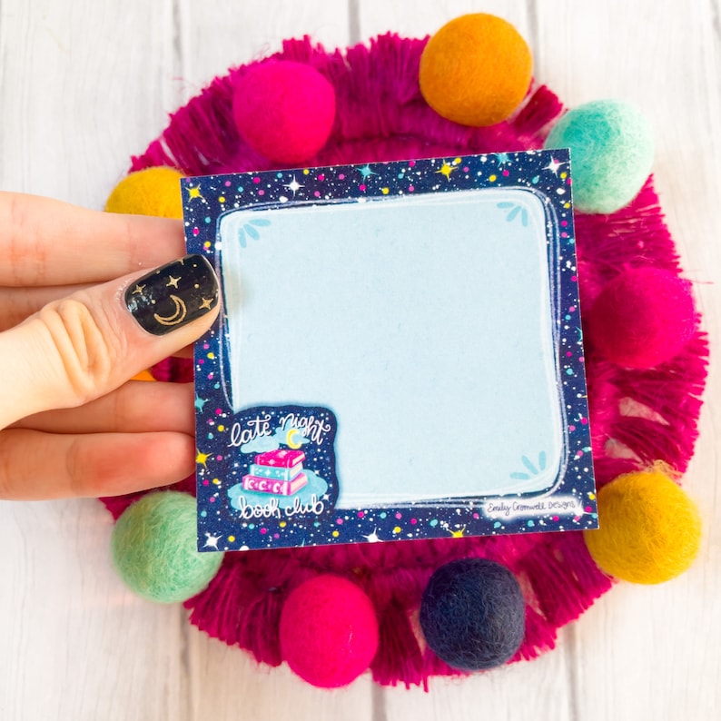 May include: A square, light blue notepad with a dark blue border featuring a starry night design. The notepad has the text "late night book club" and is surrounded by colorful felt balls and a pink fringed circle.