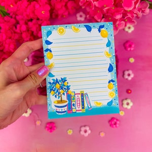 May include: A notepad with a lemon and book design. The notepad has a light blue border with lemons and leaves. The lined paper has a lemon tree in a pot and a stack of books at the bottom.