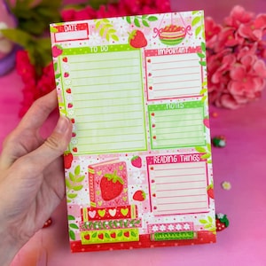 May include: A strawberry-themed notepad with sections for 'Date', 'To Do', 'Important', 'Notes', and 'Reading Things'. The notepad features a pink and green color scheme with strawberry and floral illustrations. The notepad is held in a person's hand.