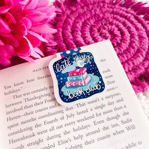 May include: A sticker with a blue background and a stack of books with stars and a crescent moon on top. The text "late night book club" is written in white on a cloud.