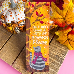 May include: A rectangular bookmark with the text "AUTUMN Leaves & LOTS OF BOOKS Please" features a gray cat wearing a pink scarf. The background is orange with a tree and falling leaves. The bookmark is on a wooden surface.