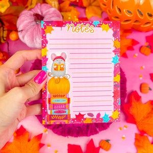 May include: A pink notepad with the word "Notes" at the top. The notepad features a cartoon mouse wearing a hat and reading a book while sitting on a pumpkin atop a stack of books. The notepad is decorated with autumn leaves.