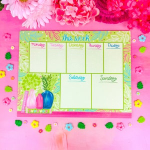 May include: A weekly planner with a green floral design and the words "this week" at the top. The days of the week are listed across the top, with space for notes below. Decorative vases are featured on the left side.