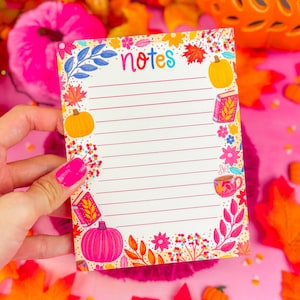 Puede incluir: Un bloc de notas rayado con la palabra "notes" en letras coloridas. El bloc de notas está decorado con ilustraciones de calabazas, libros, flores y hojas en colores otoñales. El bloc de notas está sostenido por una persona con esmalte de uñas rosa.