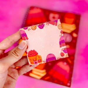 Könnte beinhalten: Ein rosa-weißer Haftnotiz mit einem leeren Bereich in der Mitte. Der Notizblock zeigt Illustrationen von Tassen, Pullovern, Äpfeln und einem Korb mit Äpfeln. Der Text "Cozy Cafe" ist auf dem Notizblock gedruckt.