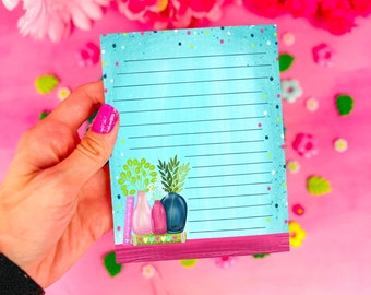 May include: A lined notepad with a teal background and a decorative illustration of vases and plants. The notepad has a pink and teal border with a watercolor effect. The notepad is held by a hand with pink nail polish.