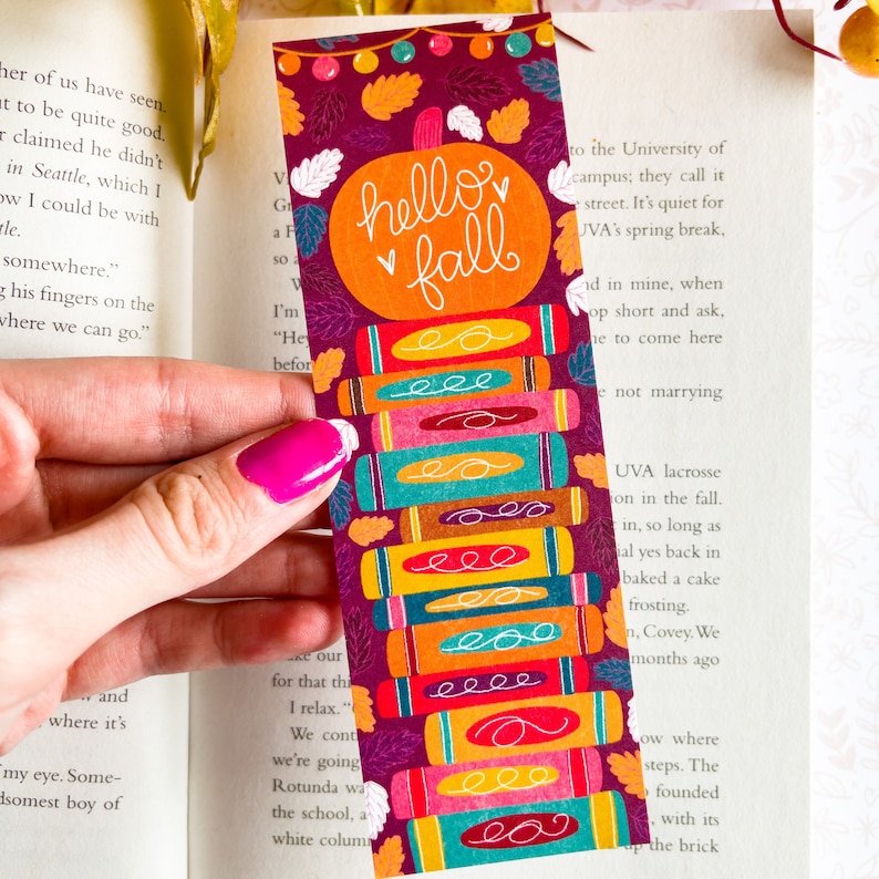 May include: A colorful bookmark with a stack of books and a pumpkin with the text "hello fall" on it. The background is a dark purple with fall leaves.