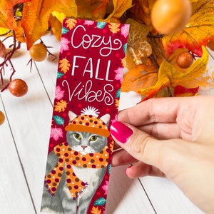 May include: A red bookmark with a gray cat wearing a brown and orange polka dot scarf and a brown and white polka dot hat. The bookmark has the text "Cozy Fall Vibes" in white lettering.