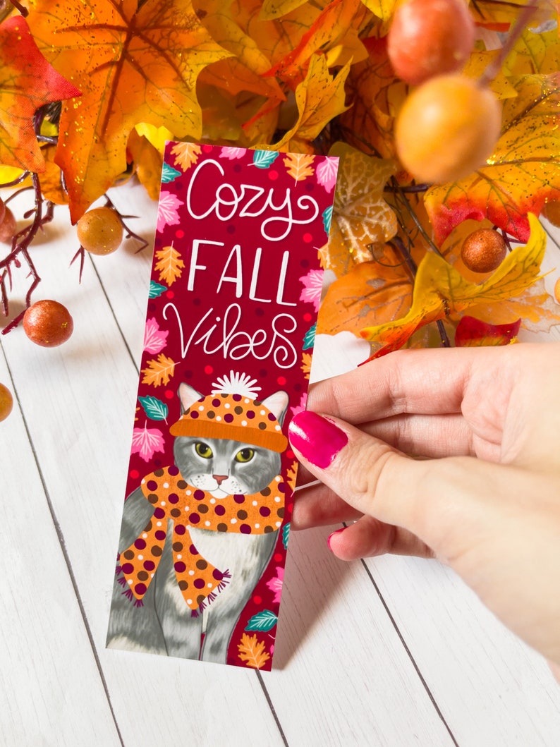 May include: A red bookmark with a gray cat wearing a brown and orange polka dot scarf and a brown and white polka dot hat. The bookmark has the text "Cozy Fall Vibes" in white lettering.