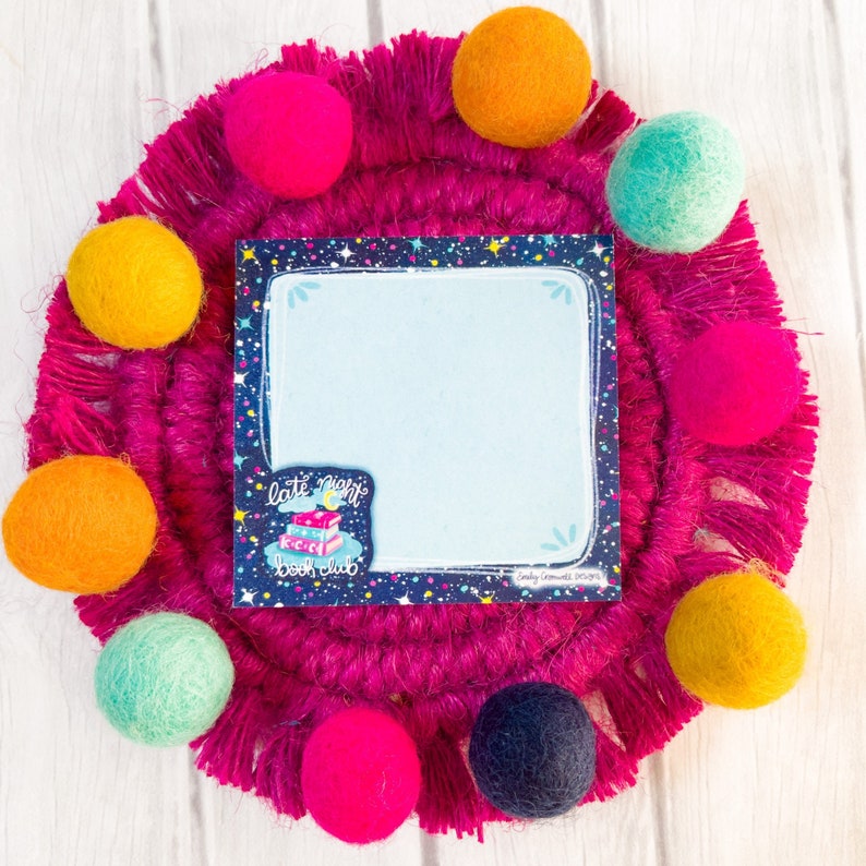 May include: A light blue notepad with a starry border and the words "late night book club" sits on a fuchsia fringed coaster. Colorful felt balls in shades of pink, yellow, orange, teal, and navy surround the notepad.