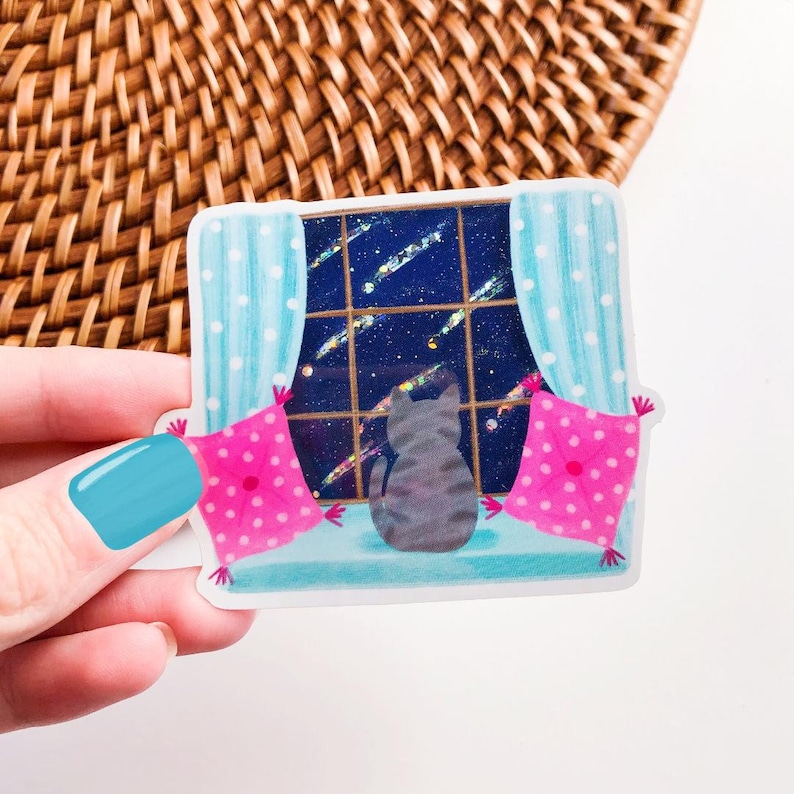 May include: A gray cat sits on a windowsill looking out at a starry night sky. The window is framed by blue curtains with white polka dots. Two pink pillows with white polka dots are on either side of the cat.