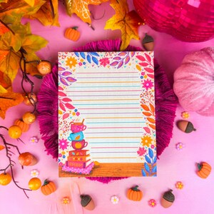 May include: A lined notepad with a floral and leaf border, featuring a stack of colorful teacups and books. The notepad is surrounded by autumn-themed decorations, including pumpkins, acorns, and fall foliage, all set against a pink background.
