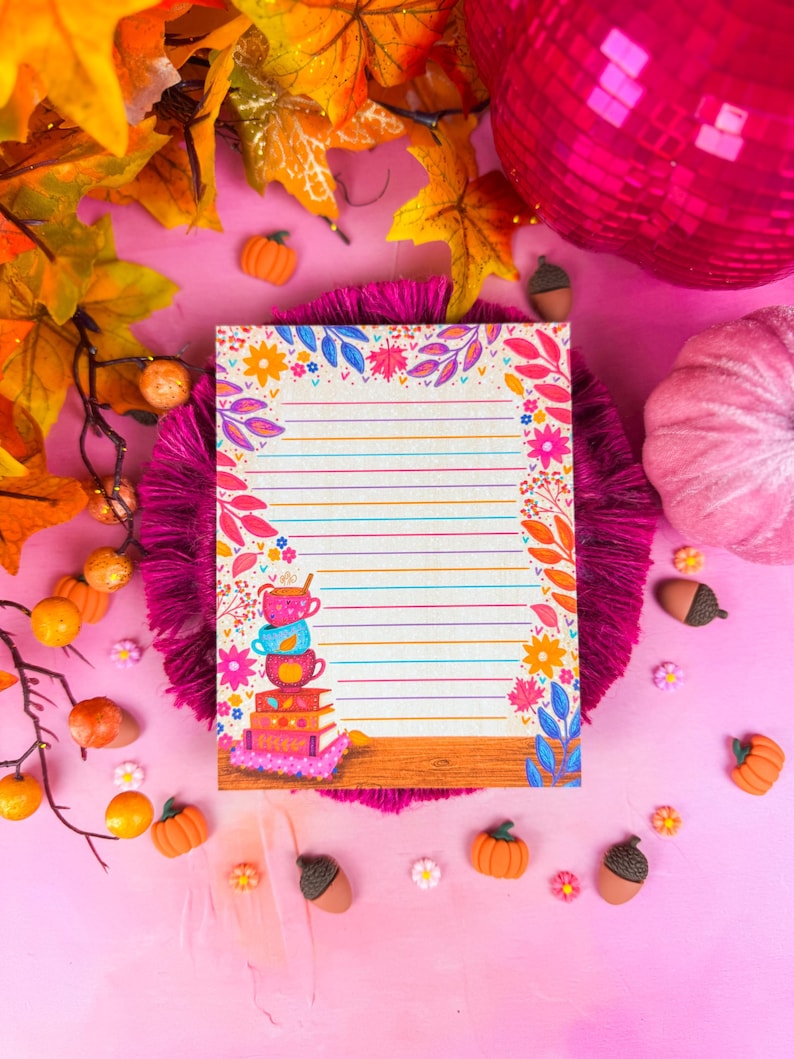 May include: A lined notepad with a floral and leaf border, featuring a stack of colorful teacups and books. The notepad is surrounded by autumn-themed decorations, including pumpkins, acorns, and fall foliage, all set against a pink background.