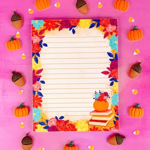May include: A lined notepad with a floral border in shades of red, orange, and yellow. The design includes a pumpkin on top of books. Decorative pumpkins, acorns, and candy corn are scattered around the notepad on a pink background.