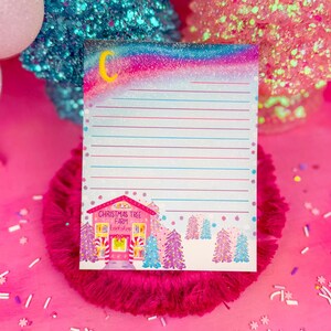 May include: A lined notepad with a whimsical Christmas theme. The top features a glittery rainbow and crescent moon. The bottom has a pink building with the text "Christmas Tree Farm Bookshop" and illustrated trees. The notepad is on a pink fluffy surface.