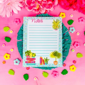 May include: A notepad with the word "Notes" in pink script, featuring illustrations of potted plants and books. The notepad is on a teal woven mat, surrounded by pink flowers and decorative accents on a pink surface.