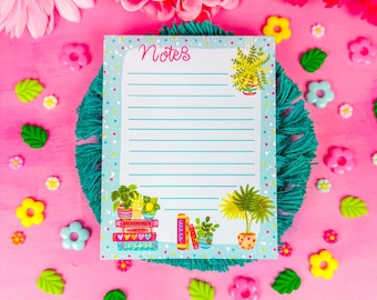 May include: A notepad with the word "Notes" in pink script, featuring illustrations of potted plants and books. The notepad is on a teal woven mat, surrounded by pink flowers and decorative accents on a pink surface.