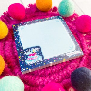 May include: A square notepad with a light blue interior and a dark blue border decorated with stars and dots. The notepad features a "late night book club" sticker with books and a crescent moon. The notepad is surrounded by colorful felt balls.