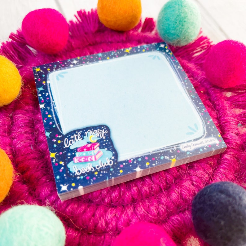 May include: A square notepad with a light blue interior and a dark blue border decorated with stars and dots. The notepad features a "late night book club" sticker with books and a crescent moon. The notepad is surrounded by colorful felt balls.