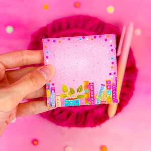 Puede incluir: Bloc de notas rosa con purpurina con un borde de libros coloridos y una planta en maceta. El bloc de notas tiene un espacio en blanco para escribir.