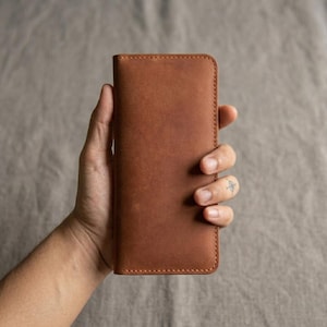 Op de afbeelding: Een bruine leren telefoonhoes met een gestikte rand. De hoes wordt in een hand gehouden tegen een neutrale achtergrond. Het leer heeft een natuurlijk, getextureerd uiterlijk.