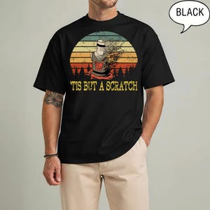 May include: A black short-sleeve t-shirt featuring a graphic of a knight's helmet dissolving into particles against a sunset background with silhouettes of trees. The text 'TIS BUT A SCRATCH' is printed in yellow below the graphic.