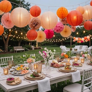 May include: An outdoor dining table set with plates of oysters, shrimp, and appetizers, decorated with floral centerpieces and illuminated by a string of hanging orange, pink, and cream paper lanterns and pom-poms above the lawn.