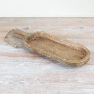 Pode incluir: Uma bandeja de servir de madeira, de formato oval alongado, com um centro raso e uma alça em uma das extremidades. Um laço grosso de corda de juta de cor natural está preso à alça, conferindo uma estética rústica à madeira castanho-clara.
