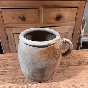 May include: A stoneware pottery jar with a handle. The jar is light grey with brown accents and a textured surface. The jar sits on a wooden table. The jar is approximately 20 cm in height.