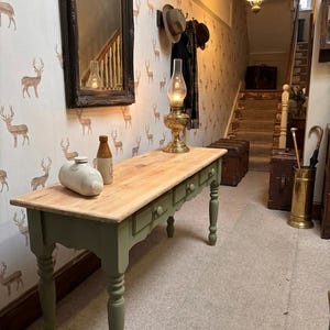 May include: A long wooden console table with a natural wood top and sage green base featuring three drawers with round knobs. On the table sits a brass oil lamp, a white ceramic bottle, and a brown glass bottle. The table has four turned legs.