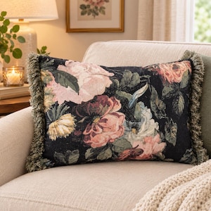 May include: A rectangular throw cushion featuring a dark black background with a woven floral pattern in shades of pink, cream, and green. The cushion is finished with a textured, olive-green fringe trim along the two shorter vertical edges.