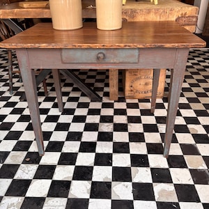 May include: A rectangular wooden table with a natural brown top and a distressed blue-grey painted base. The table features four tapered legs and a single central drawer with a small, round wooden knob centred on the front panel of the drawer.